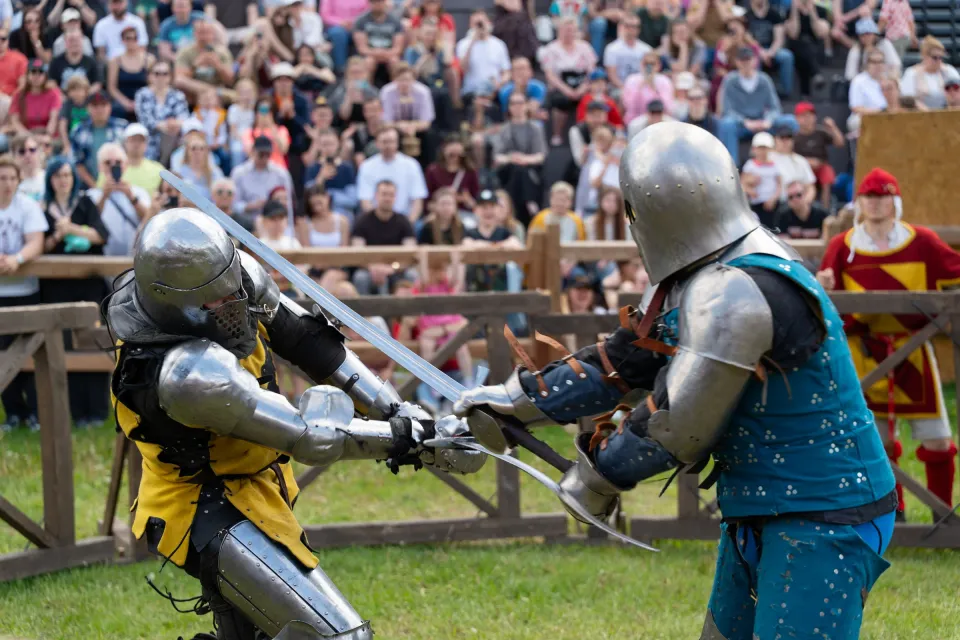 Dwaj rycerze w zbrojach walczą na miecze na polu turniejowym przed licznym tłumem
