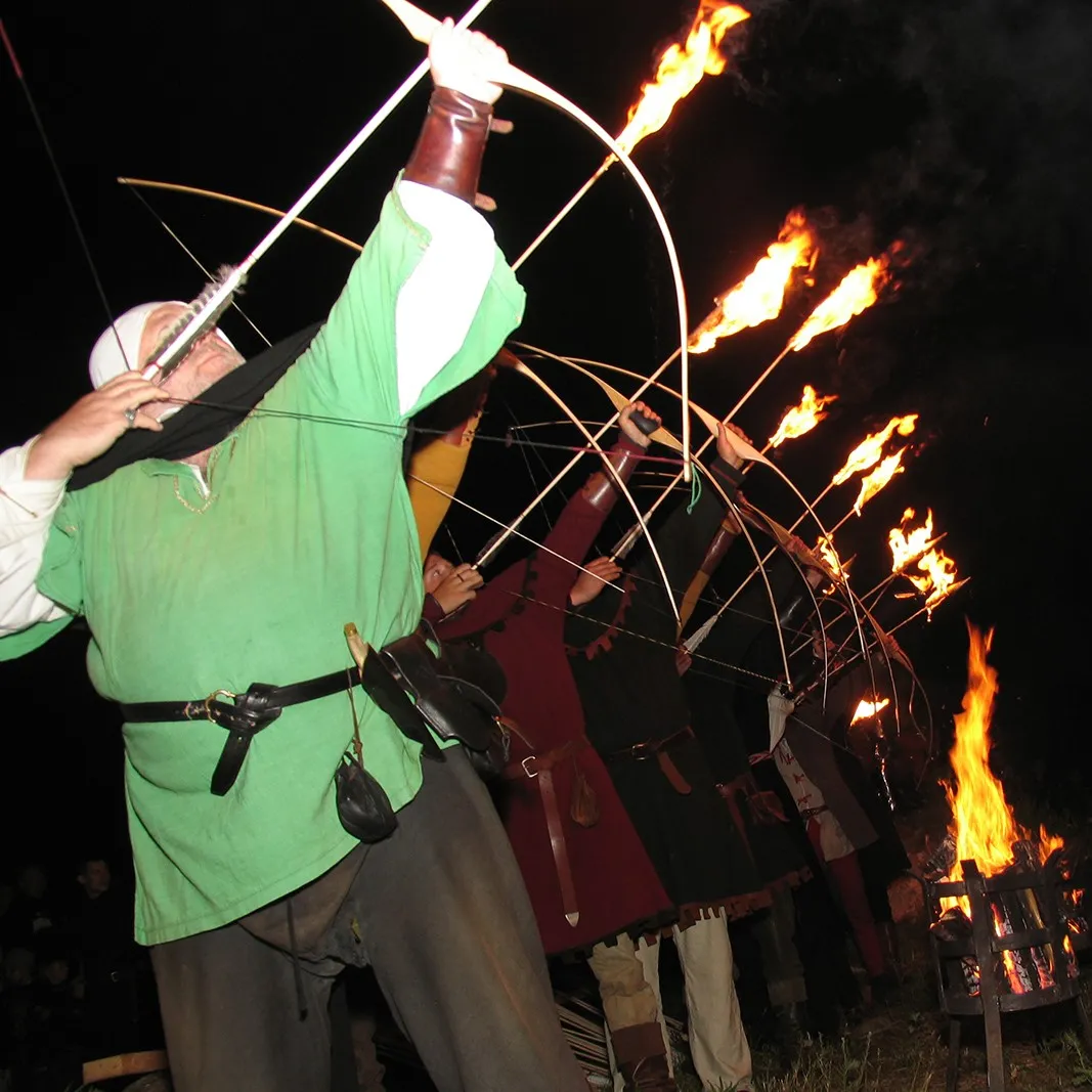 An archer in a green tunic shoots a flaming arrow beside a bonfire