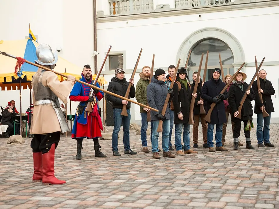 Edukatorius kostiumu moko dalyvių grupę laikyti medines ietis pilies kieme
