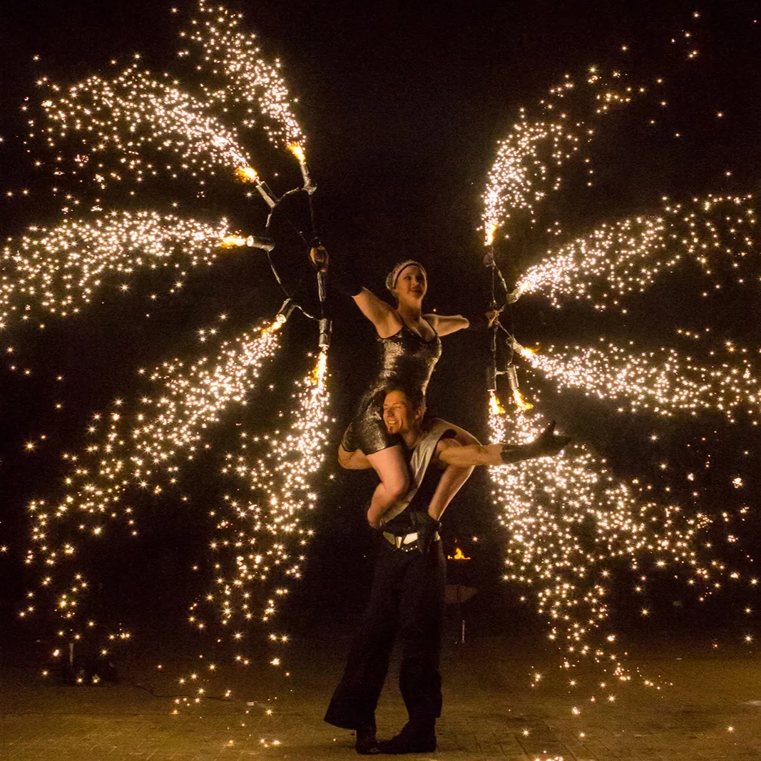 A performer lifted on a partner's shoulders between wings of sparks