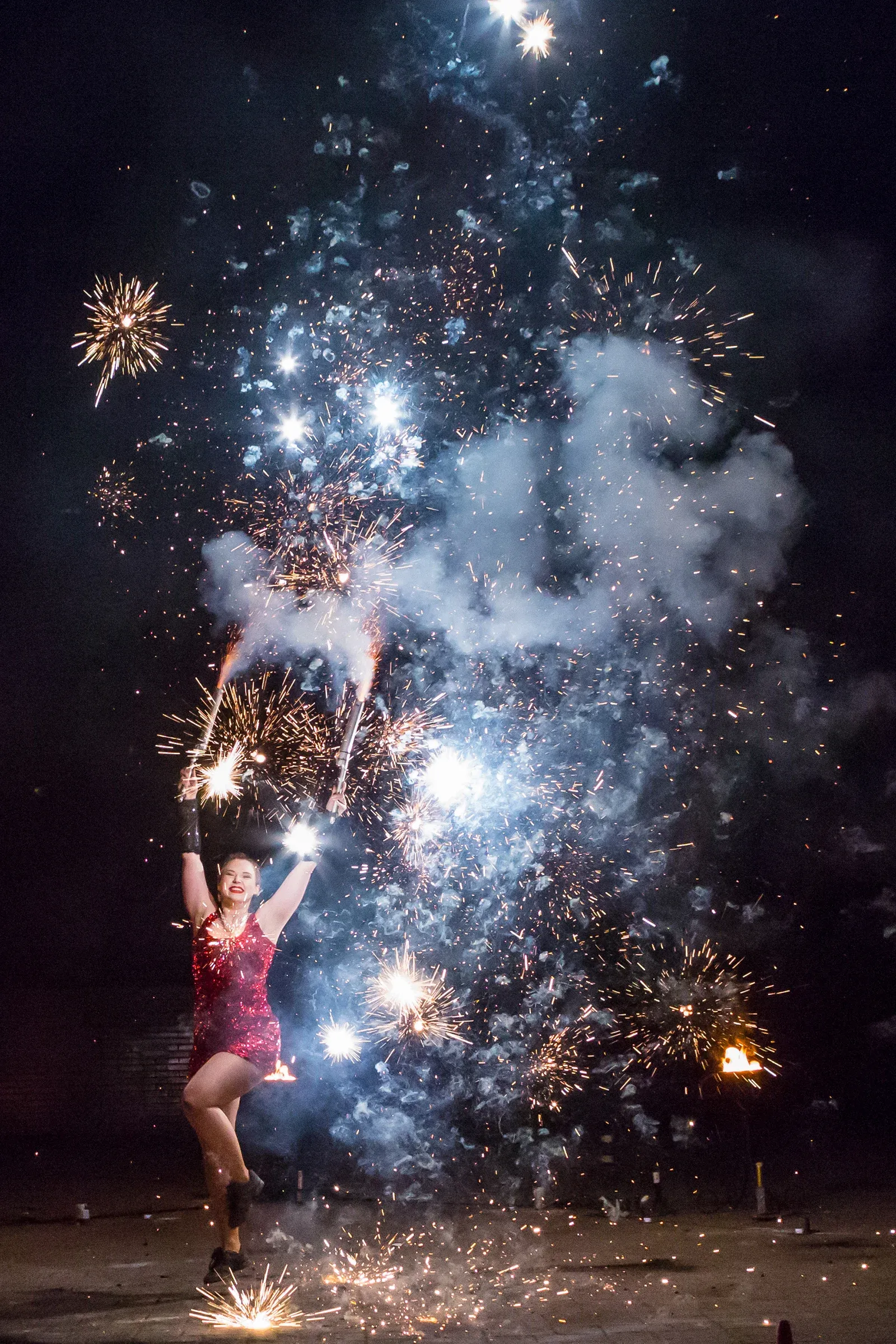 A performer in a red dress lifts two burning wands beneath bursting fireworks