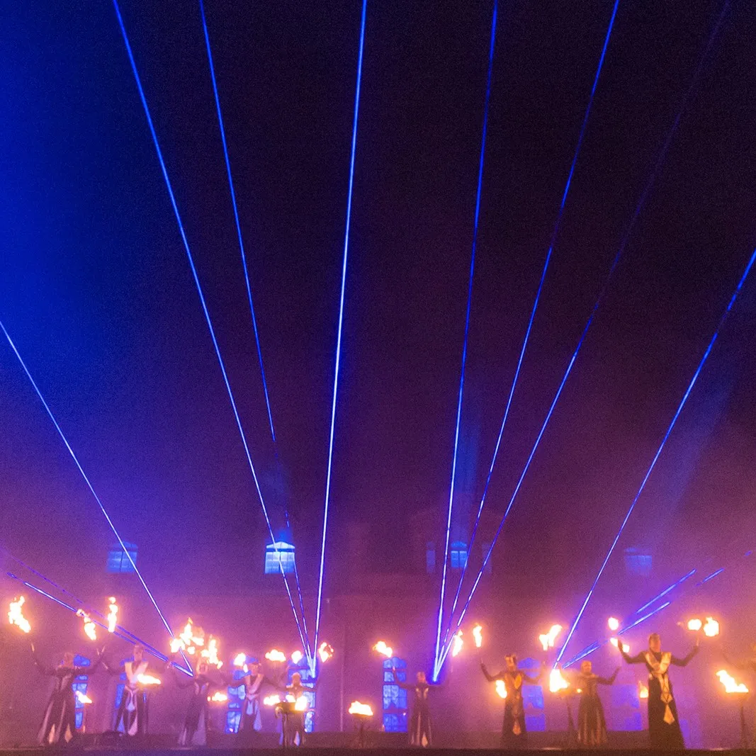 Performers with flaming tools beneath a web of laser beams