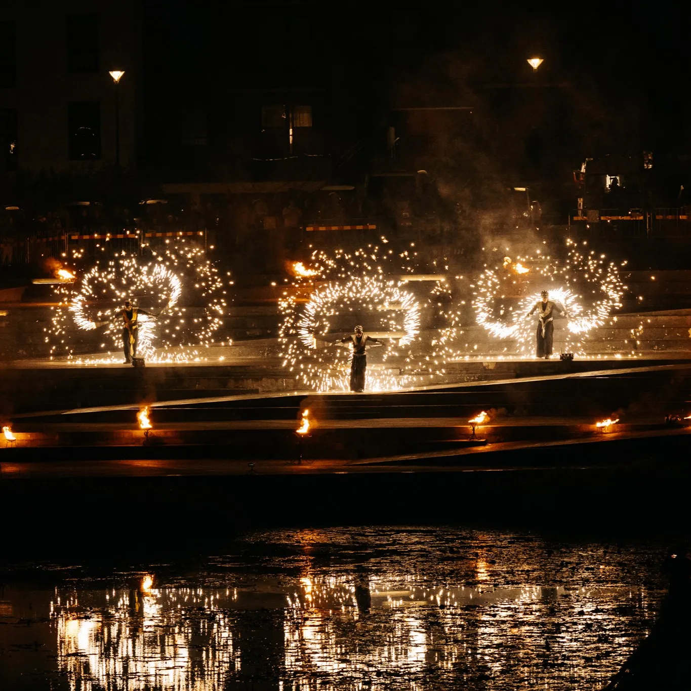Performers spin spirals of fire on riverside steps above the water