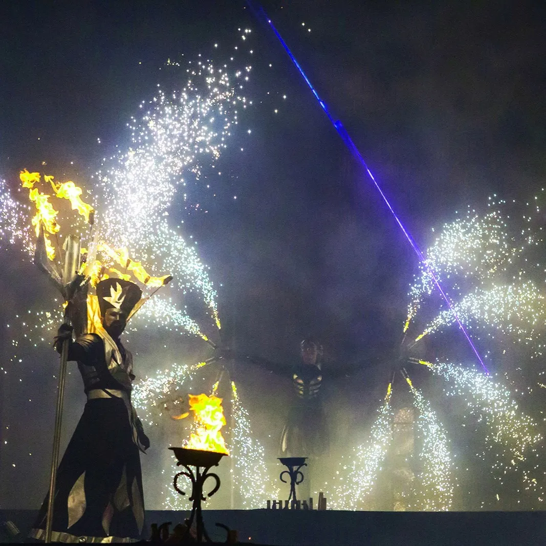 A performer holds a blazing trident against fireworks and a laser beam