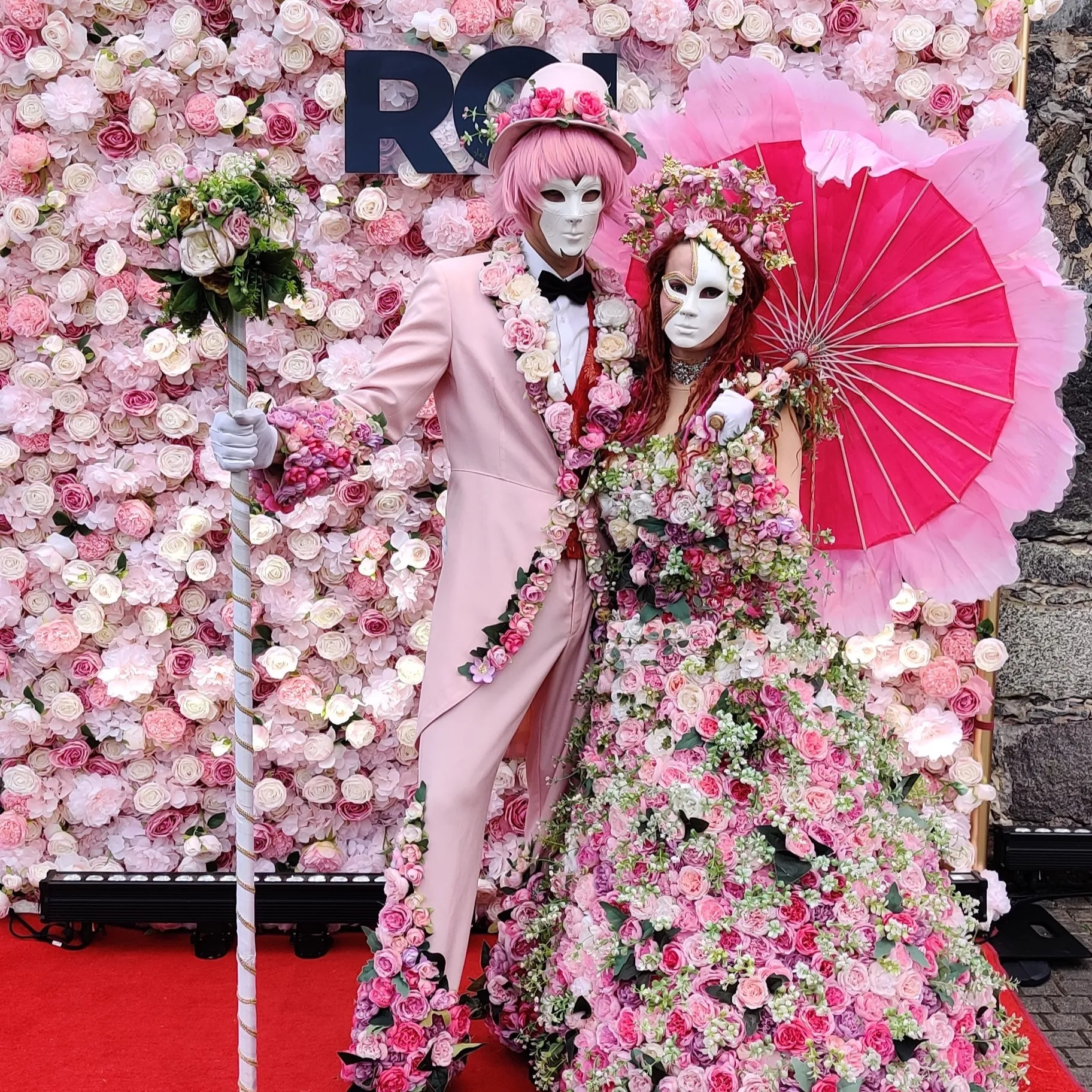 Flower people couple posing against a flower wall with a floral staff and a fan