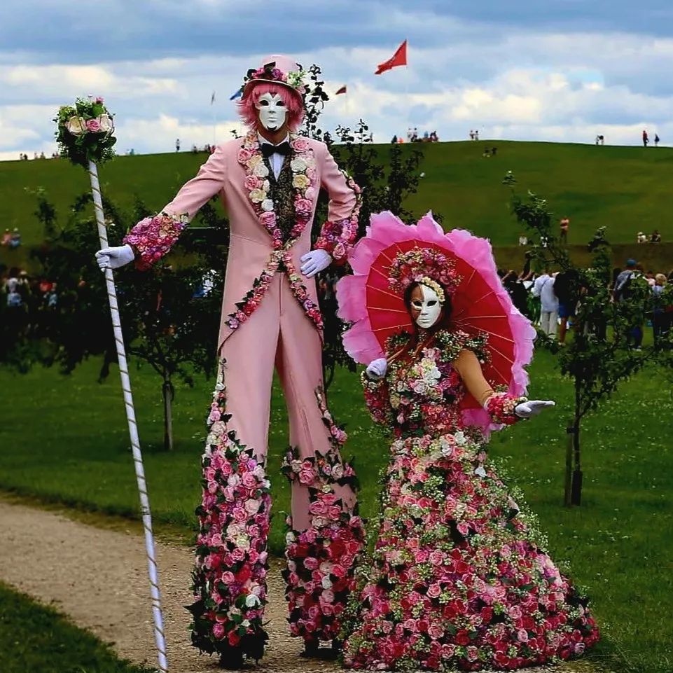 Flower characters on stilts with a large red parasol on a green hillside