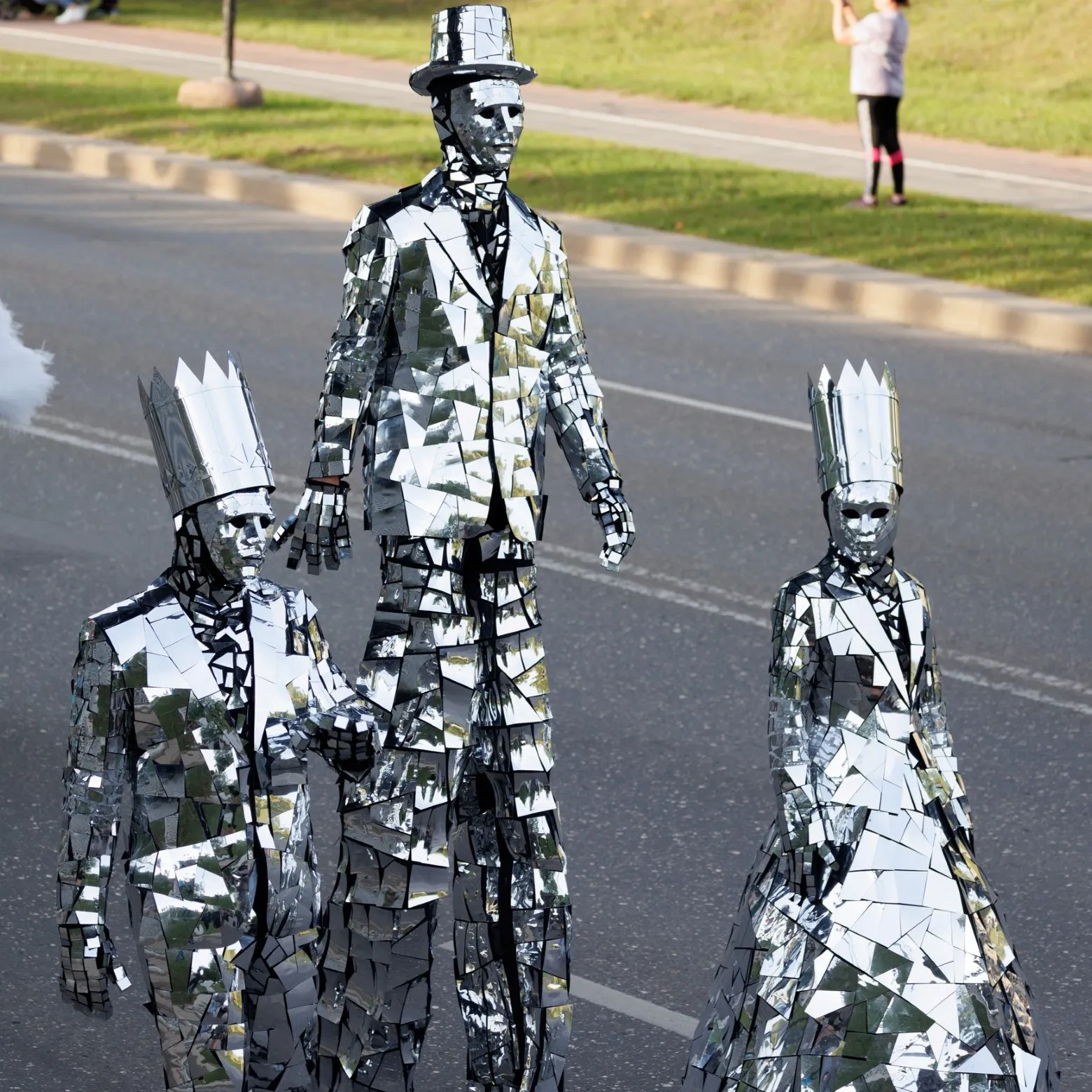 Three mirror-costumed stilt walkers walk down a city street