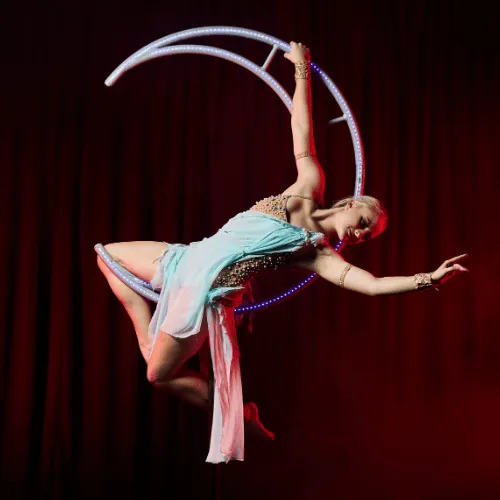 An acrobat in a mint costume inside a glowing Moon apparatus against a red backdrop