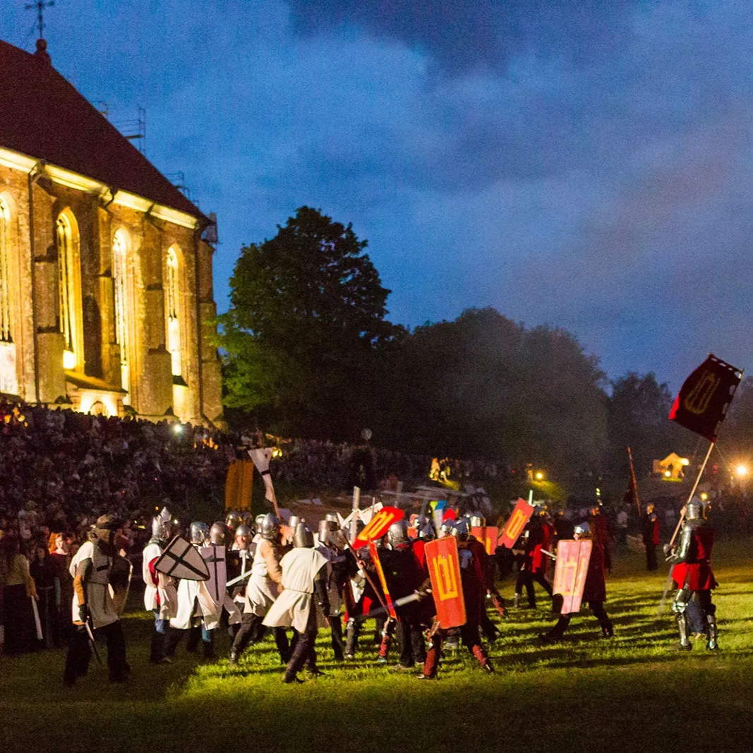 A theatrical night battle scene of knights beside a lit-up church