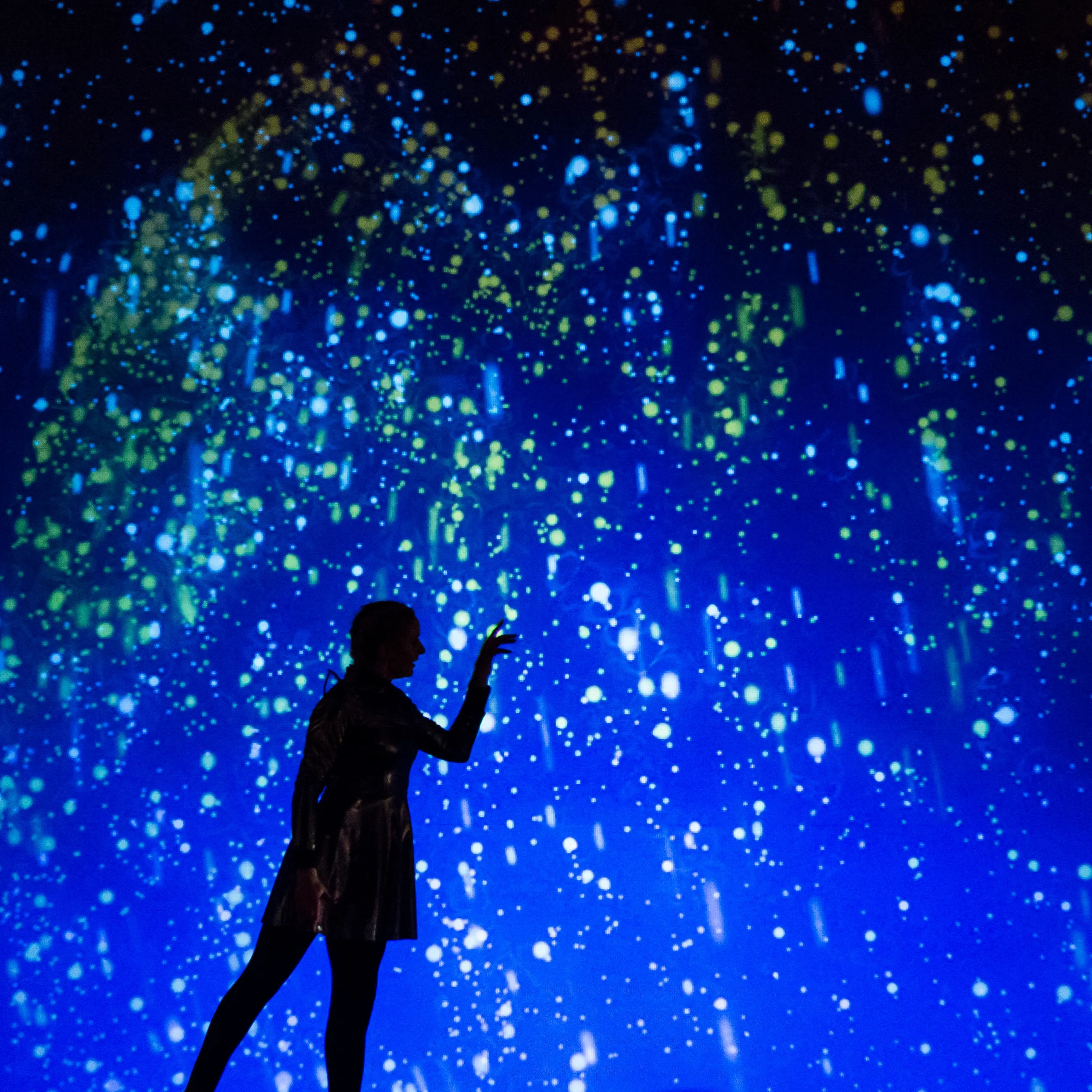 A woman reaches toward drifting light particles in a blue-green projection