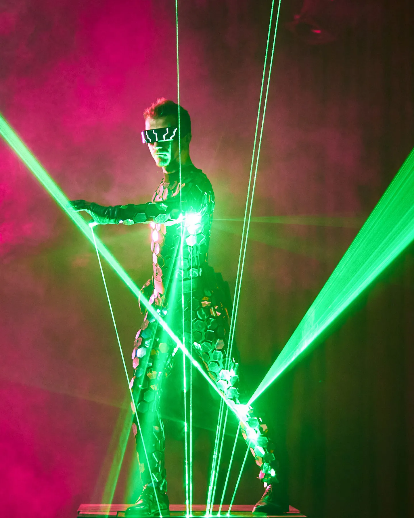A performer in a mirrored costume with arms spread inside green laser beams