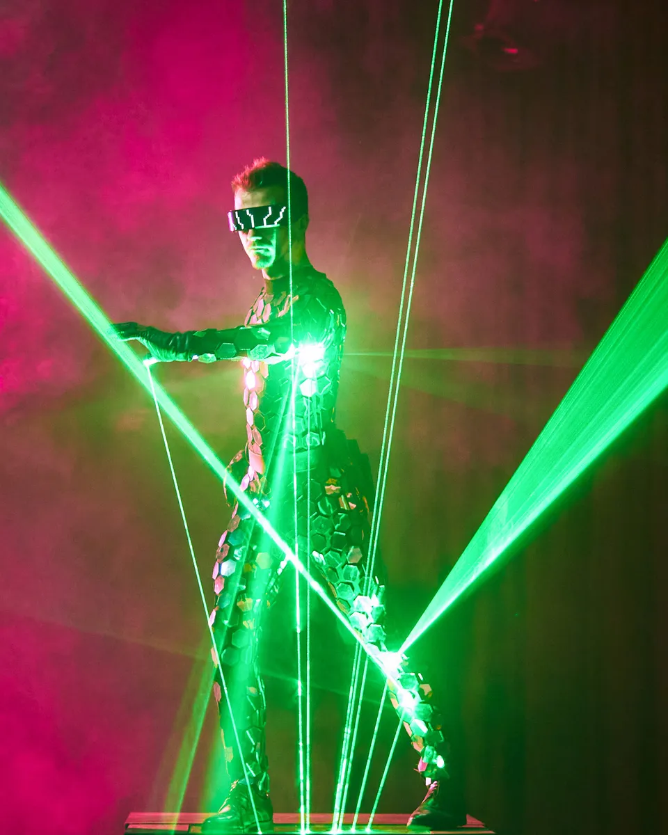 A performer in a mirrored costume with arms spread inside green laser beams