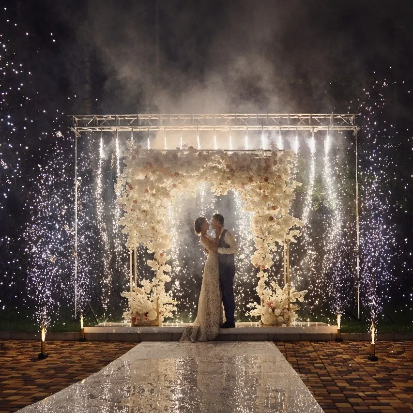 Newlyweds embrace under a floral arch with spark fountains around them