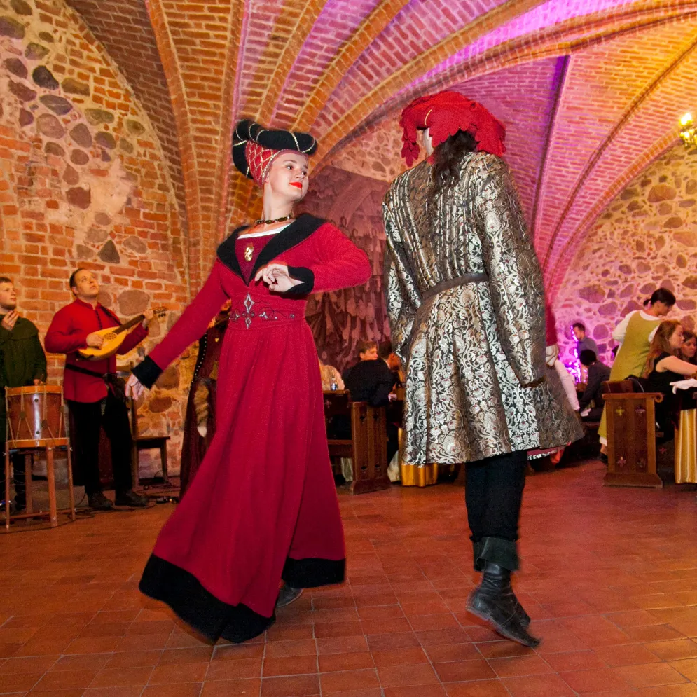 A couple dances in a vaulted medieval hall as a musician plays nearby