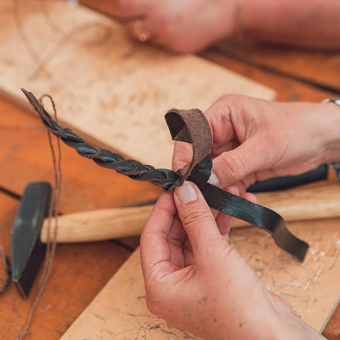 Hands braid a leather bracelet beside a wooden mallet