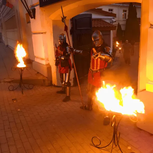 Knights carrying a spear walk past lit fire stands on an evening street