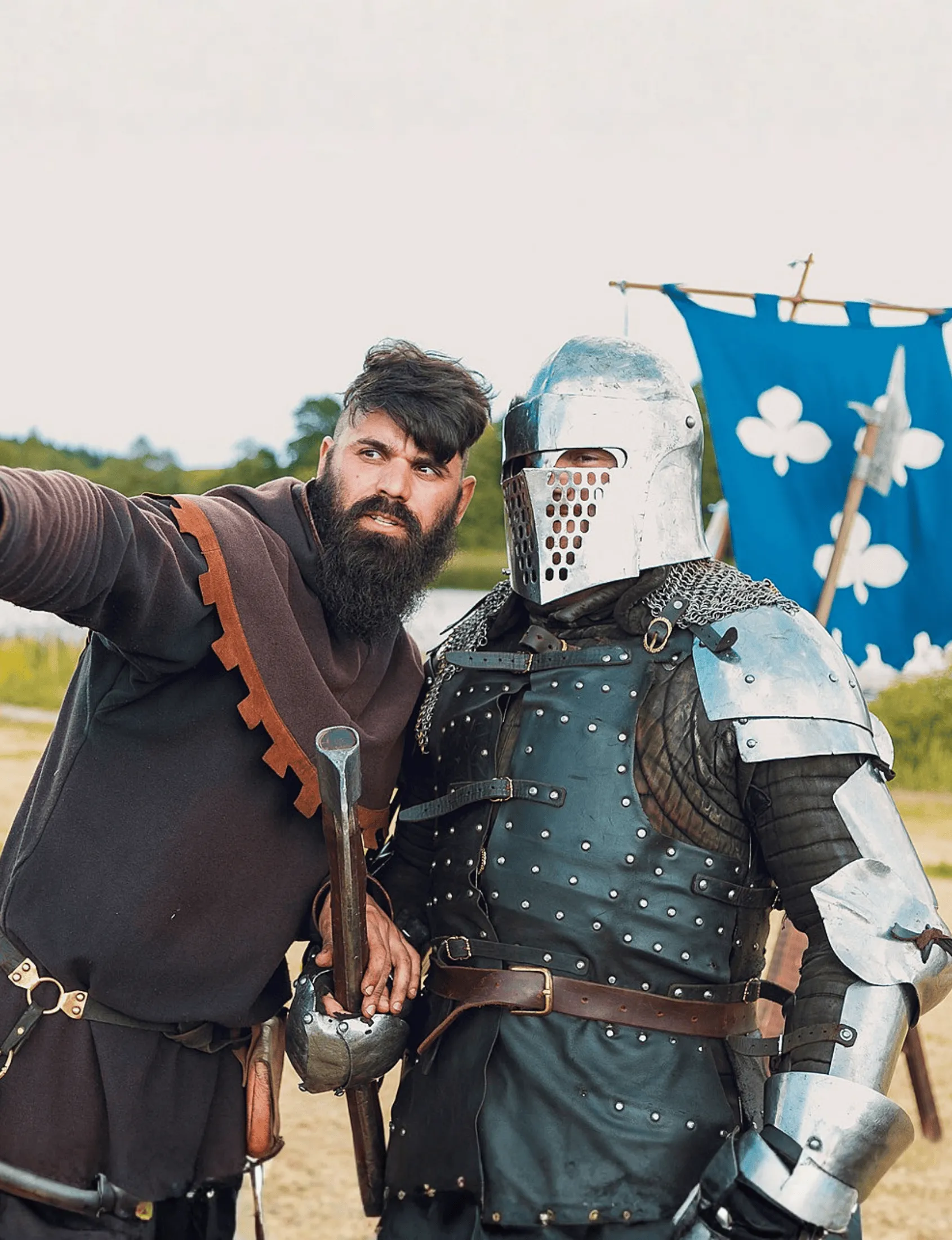 A man in full knight's armour beside a guide, with a colourful banner behind them