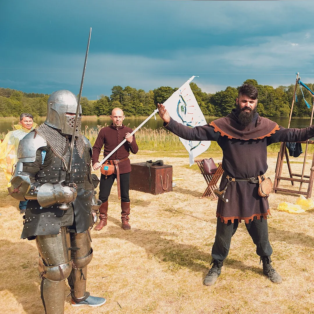 A knight in full armour stands beside a man holding a flag near a lake