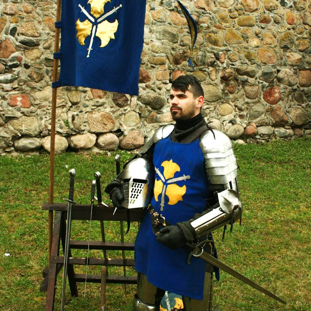 A knight stands by a blue banner and sword rack near a stone wall