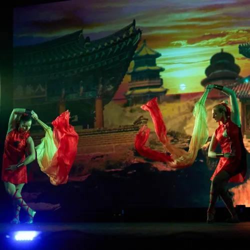 A dancer poses with a red-and-yellow fan against a sunset temple backdrop