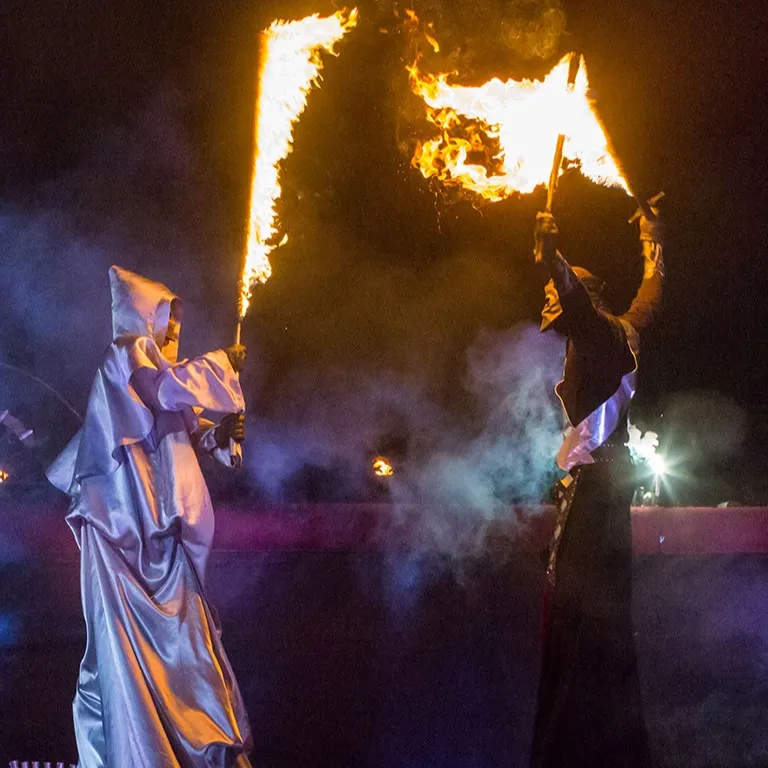 Stilt walkers with blazing flags perform amid night-time smoke