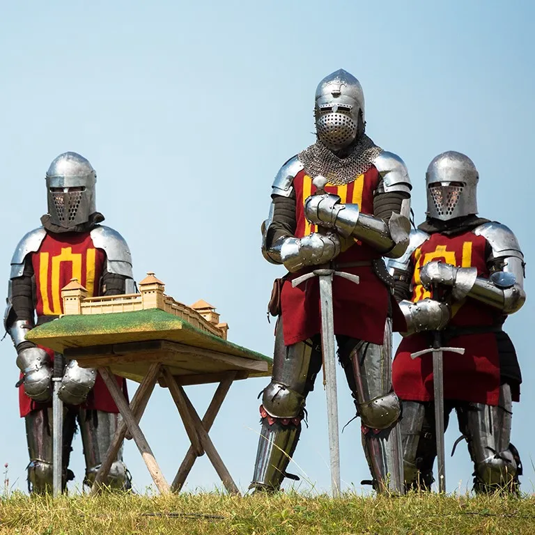 Three knights stand outdoors beside a wooden castle model