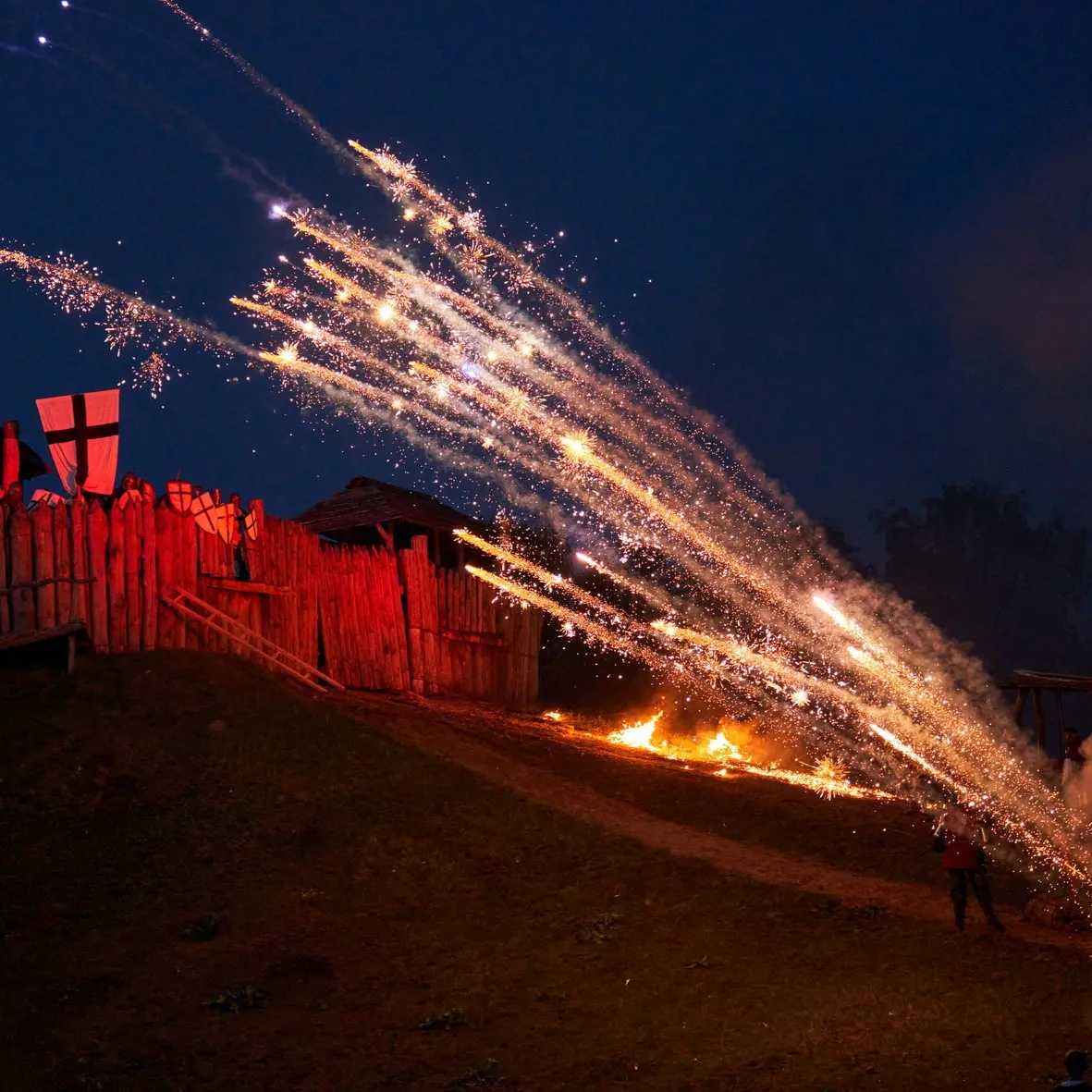 Fire fireworks shoot up over a wooden fort palisade at night