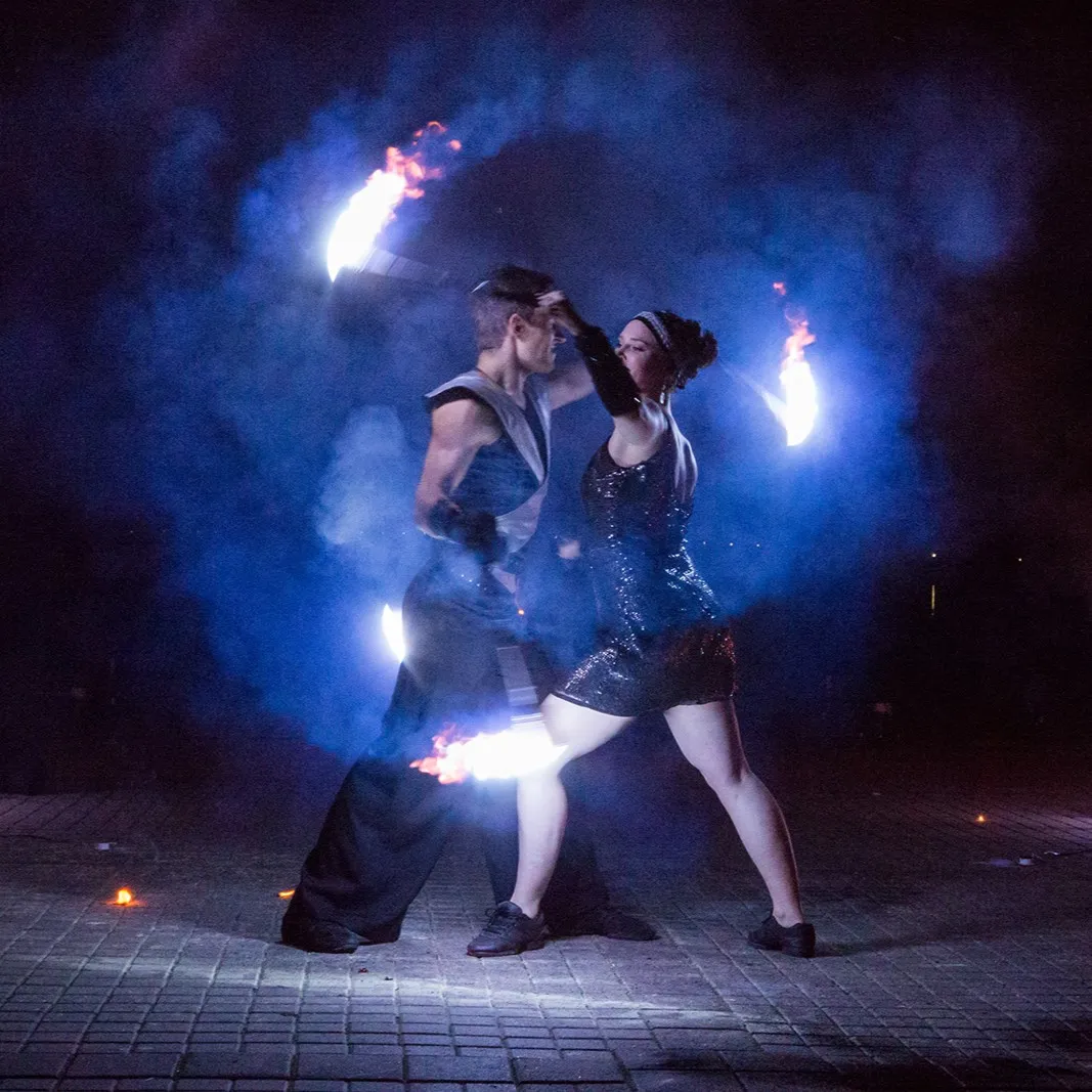 A duo dances with flaming tools amid blue smoke