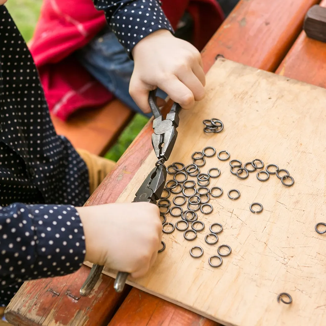 A child's hands use pliers to link metal chainmail rings on a board