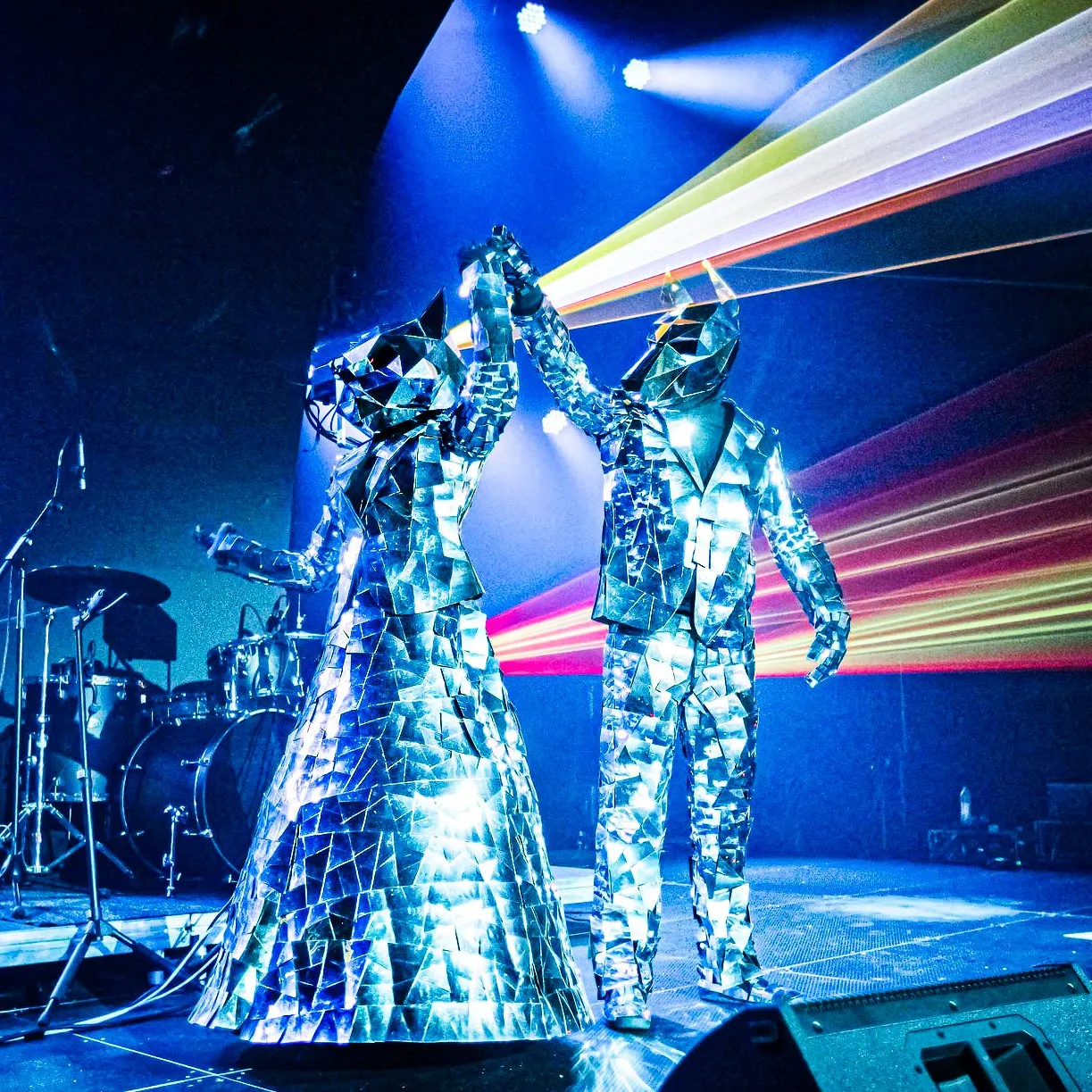 Two mirror-mosaic characters hold raised hands on stage amid laser light beams
