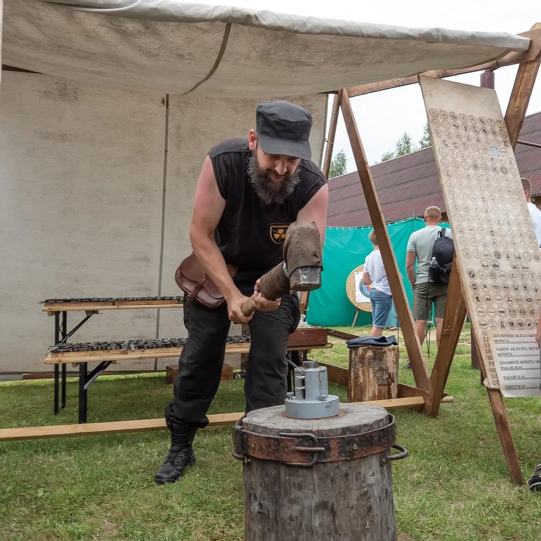 A man strikes a coin at an outdoor fair beside a board of coin-design samples