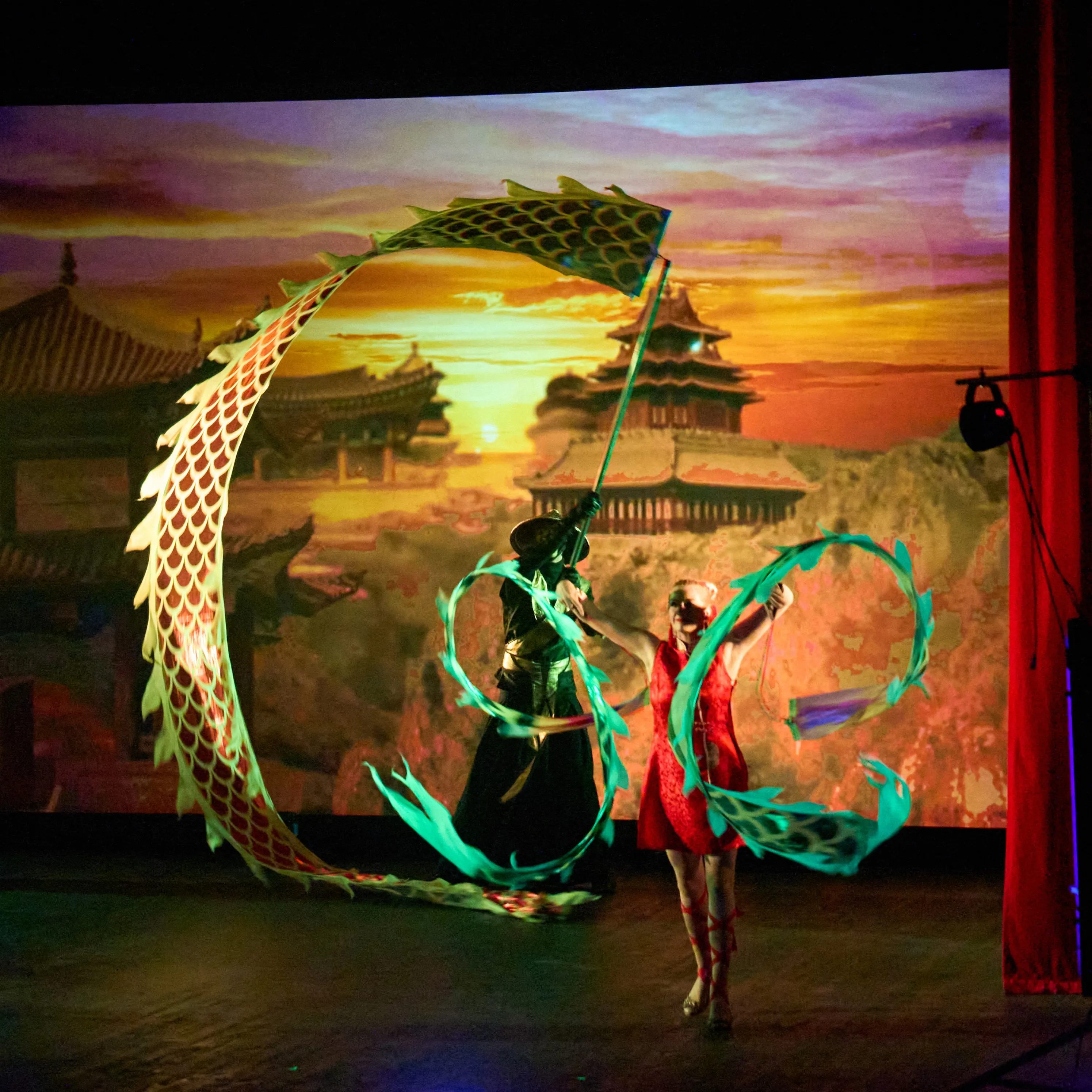 A green Chinese dragon prop beside a dancer waving red fan streamers on stage