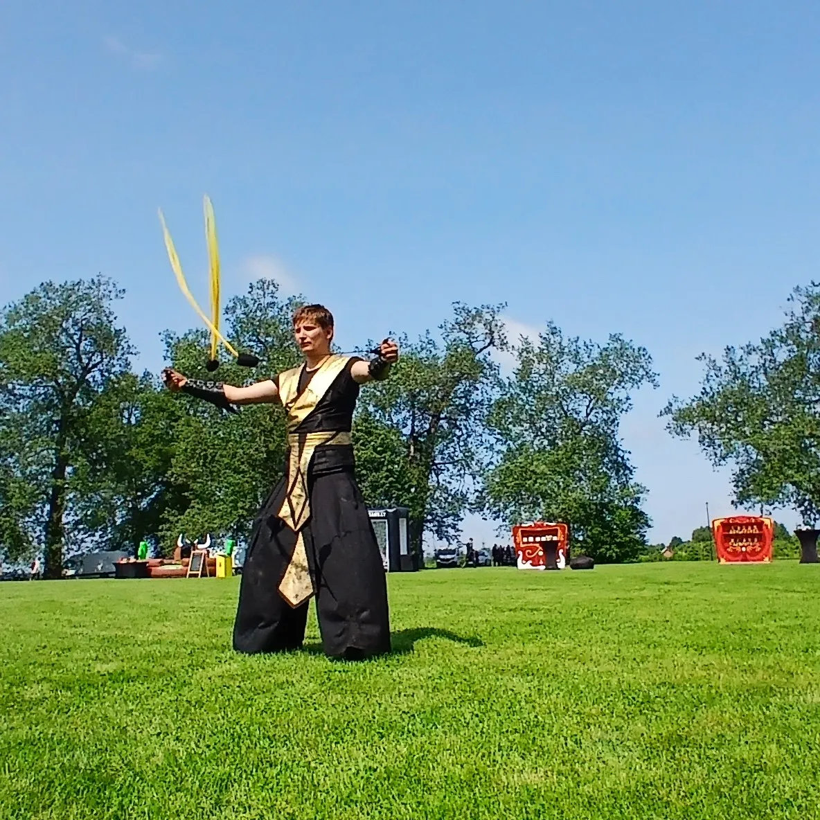 A performer in a black and gold costume juggles sticks on an open lawn